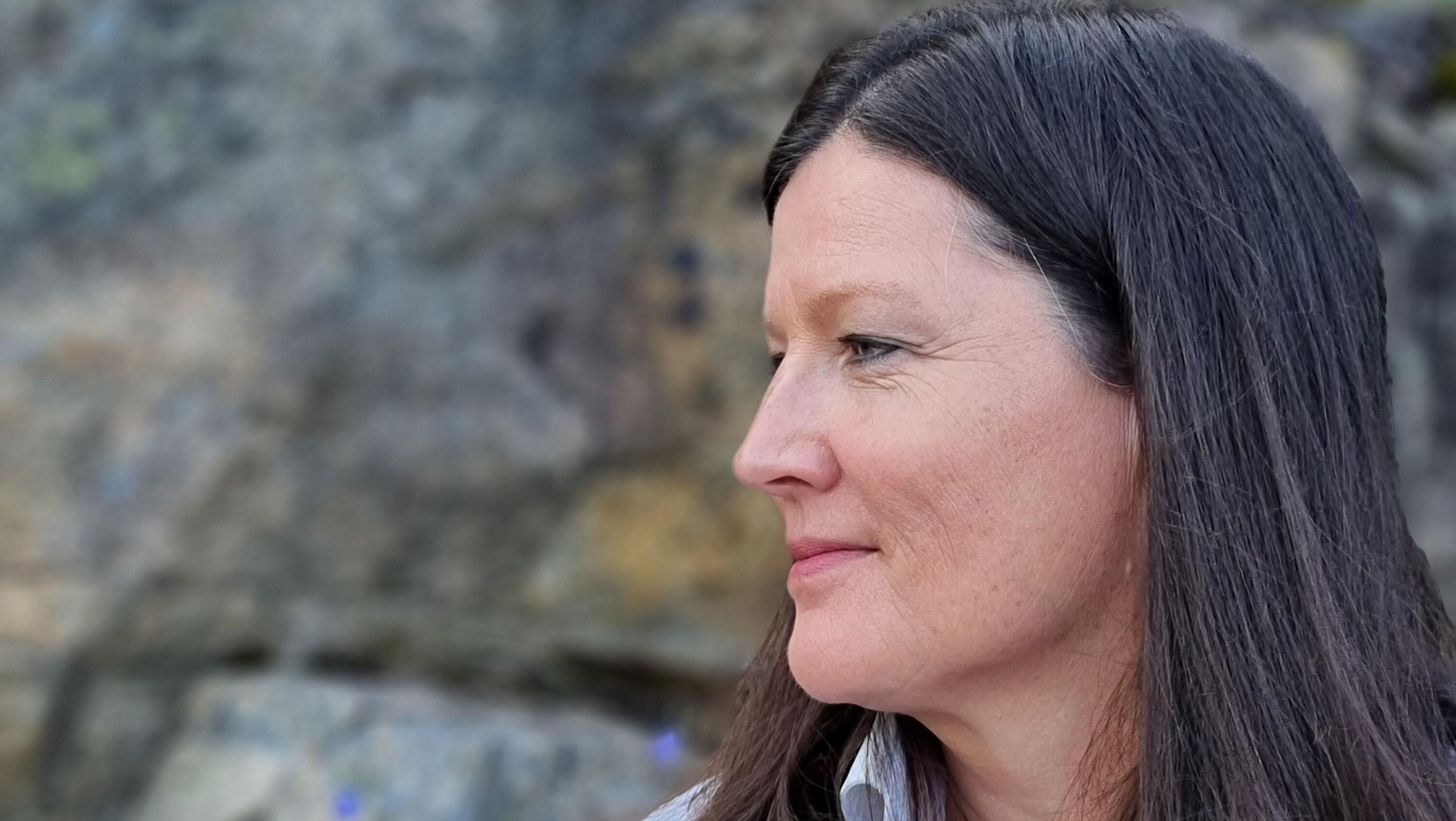 Nærbilde av kvinne i profil mot steinvegg, med naturlig sminke og mykt lys over ansiktet