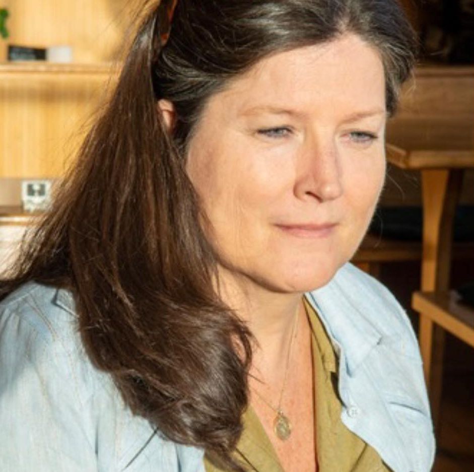 Kvinne med langt brunt hår sitter innendørs ved bord, iført lys skjorte og med rolig, konsentrert uttrykk