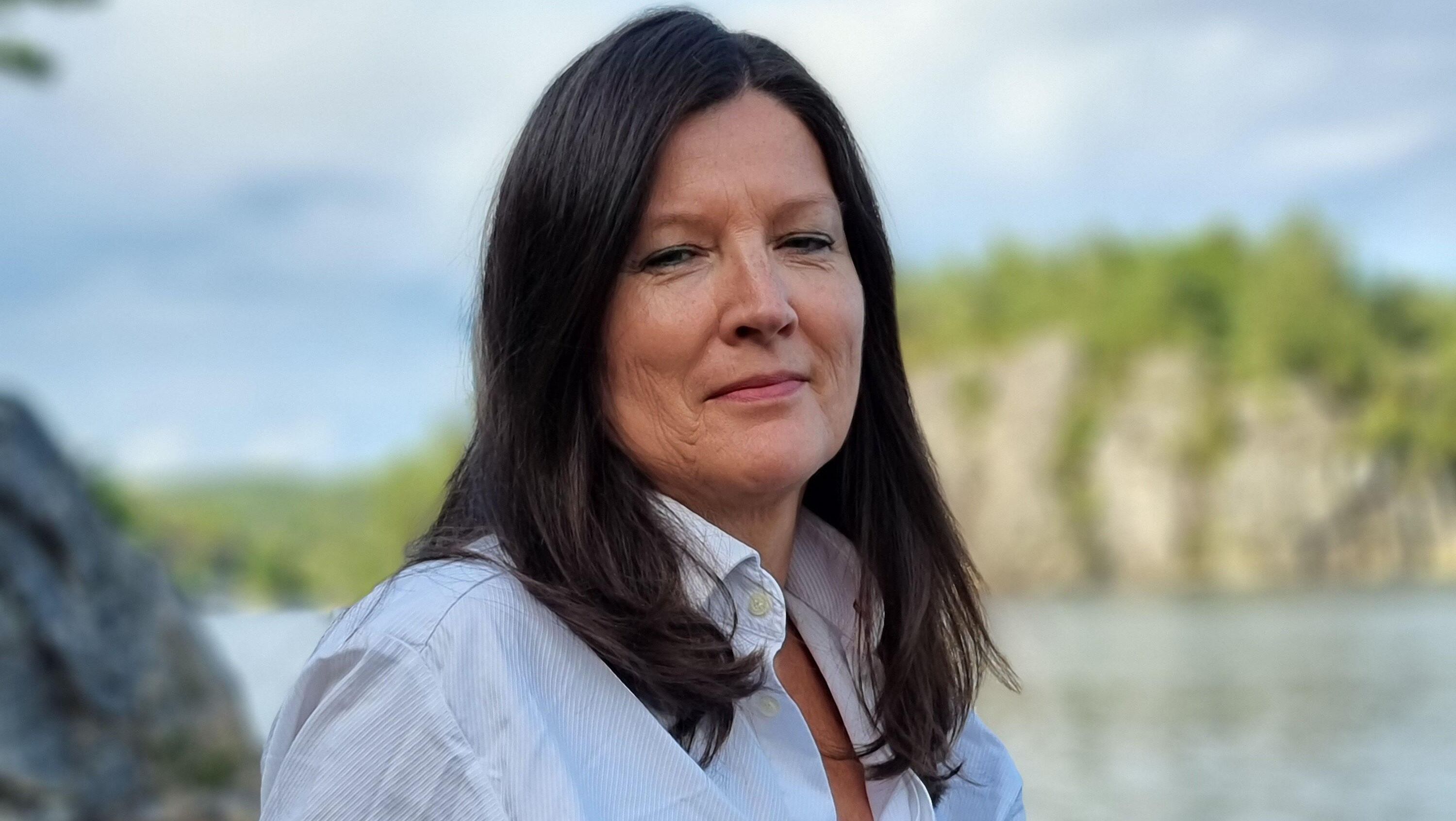 Portrett av kvinne i hvit skjorte ved vann og fjell, med rolig uttrykk og natur i bakgrunnen