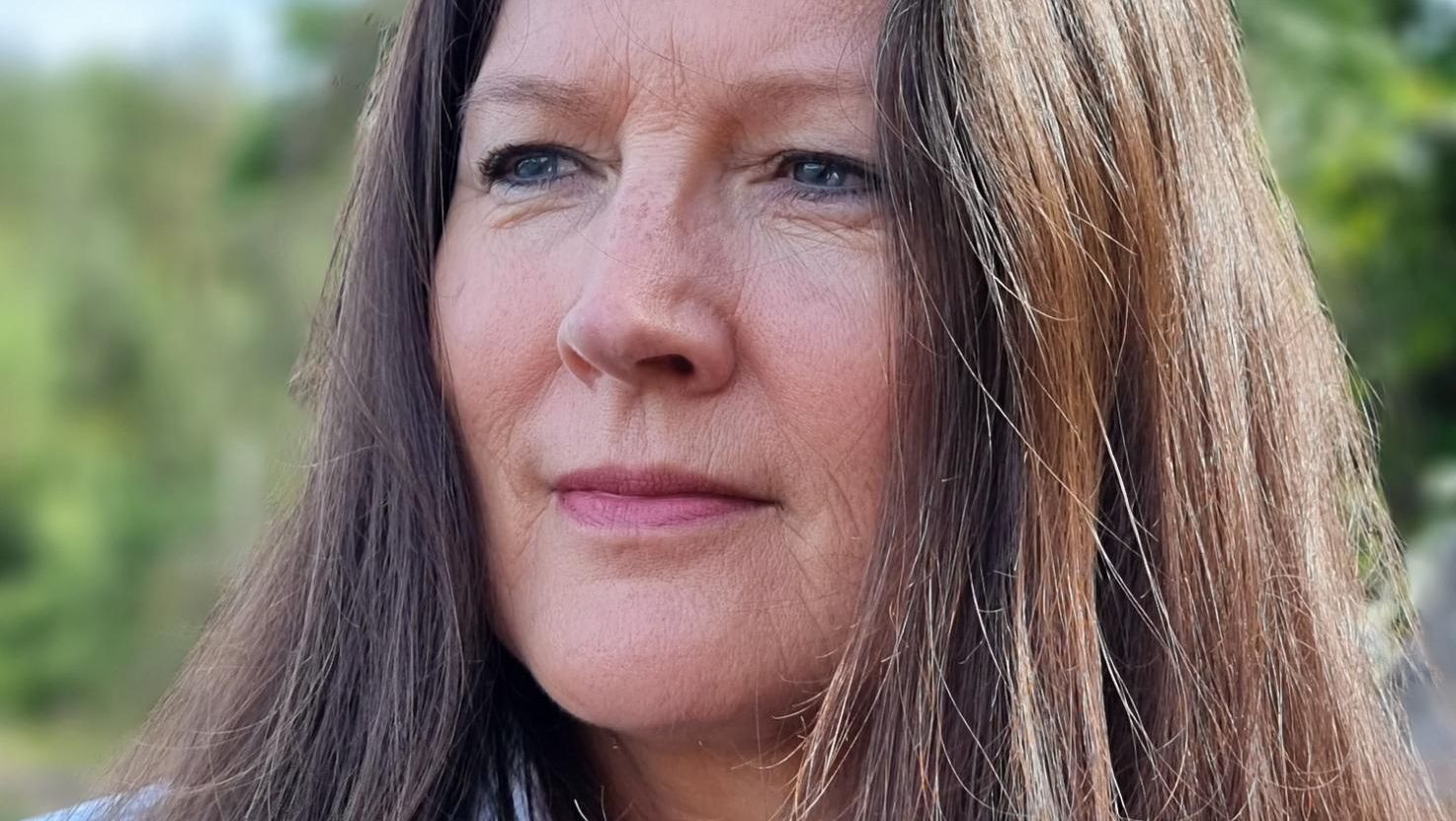 Portrett av kvinne med langt brunt hår utendørs, med grønn natur og mykt dagslys rundt seg